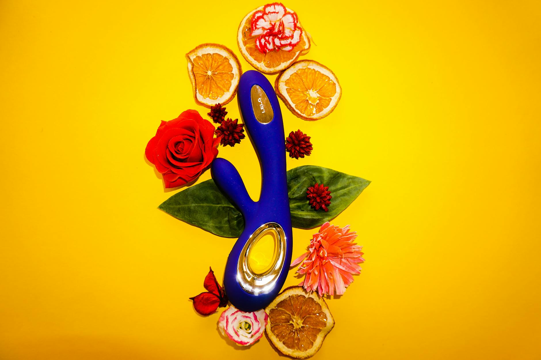 A blue vibrator surrounded by flowers and orange slices on a bright yellow background.
